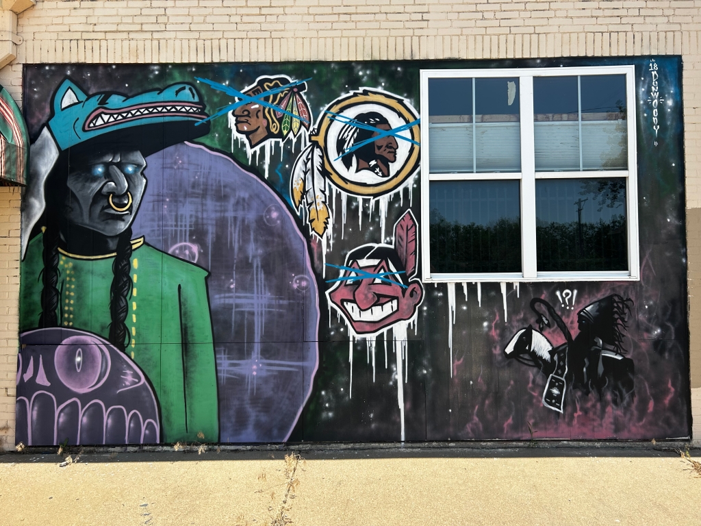A mural on a black background showing different mascots with Native American caricatures crossed out. a man with an animal head mask on his head looks toward them with glowing blue eyes. 