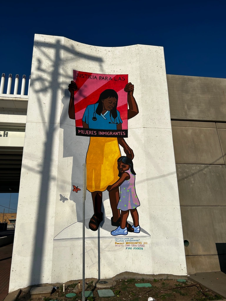 In a mural, a person holds a poster in front of their face reading "Justicia para las mujeres inmigrantes." A child hugs their leg.